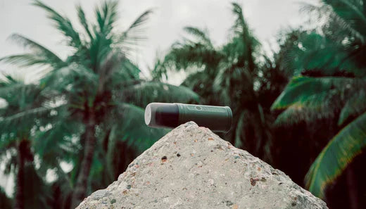 Tragbarer Kaffeezubereiter liegt auf Felsbrocken vor Palmen; Outdoor-Produktfoto mit tropischer Landschaft und robustem Design