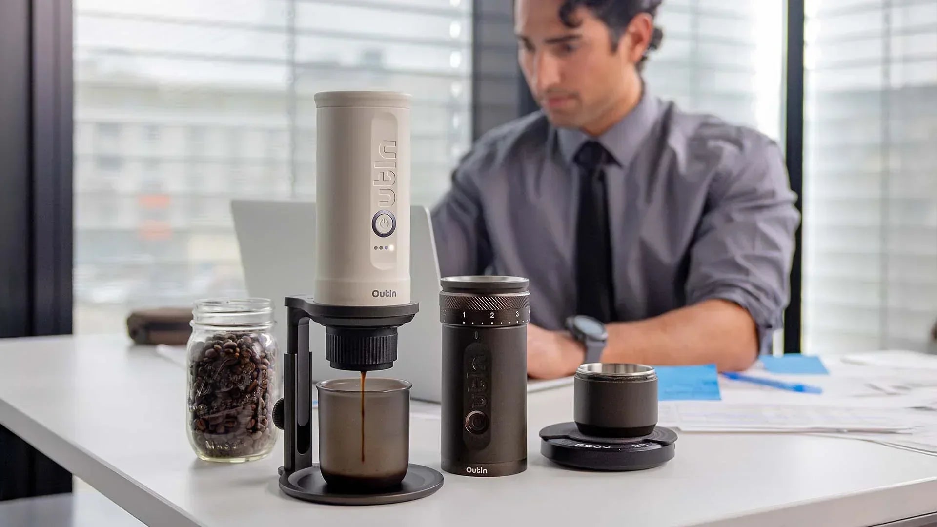 Ein Geschäftsmann brüht frischen Kaffee an seinem Schreibtisch mit einer tragbaren Espressomaschine und einer Kaffeemühle von Outin – ein professionelles Kaffee-Setup für den Arbeitsplatz.