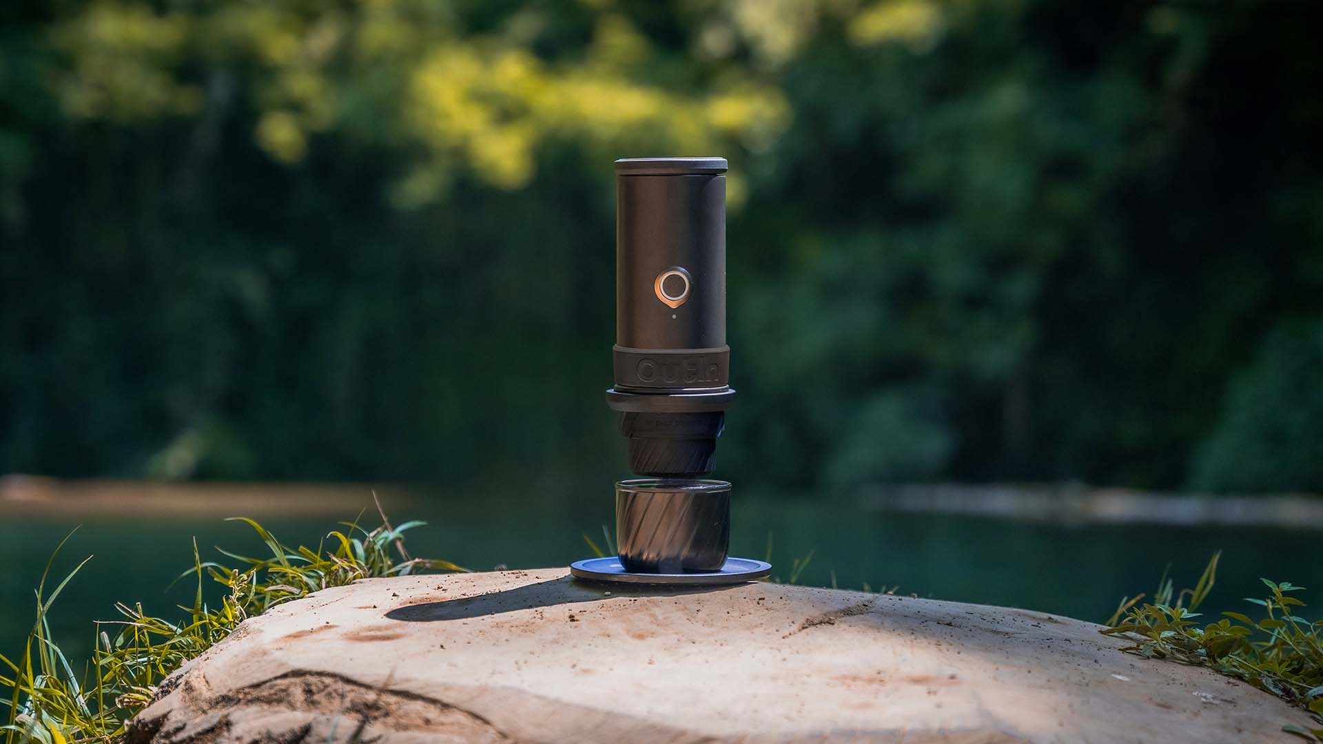 Eine tragbare Espressomaschine brüht einen Espresso in eine Tasse, die auf einem hölzernen Baumstumpf am Seeufer steht.