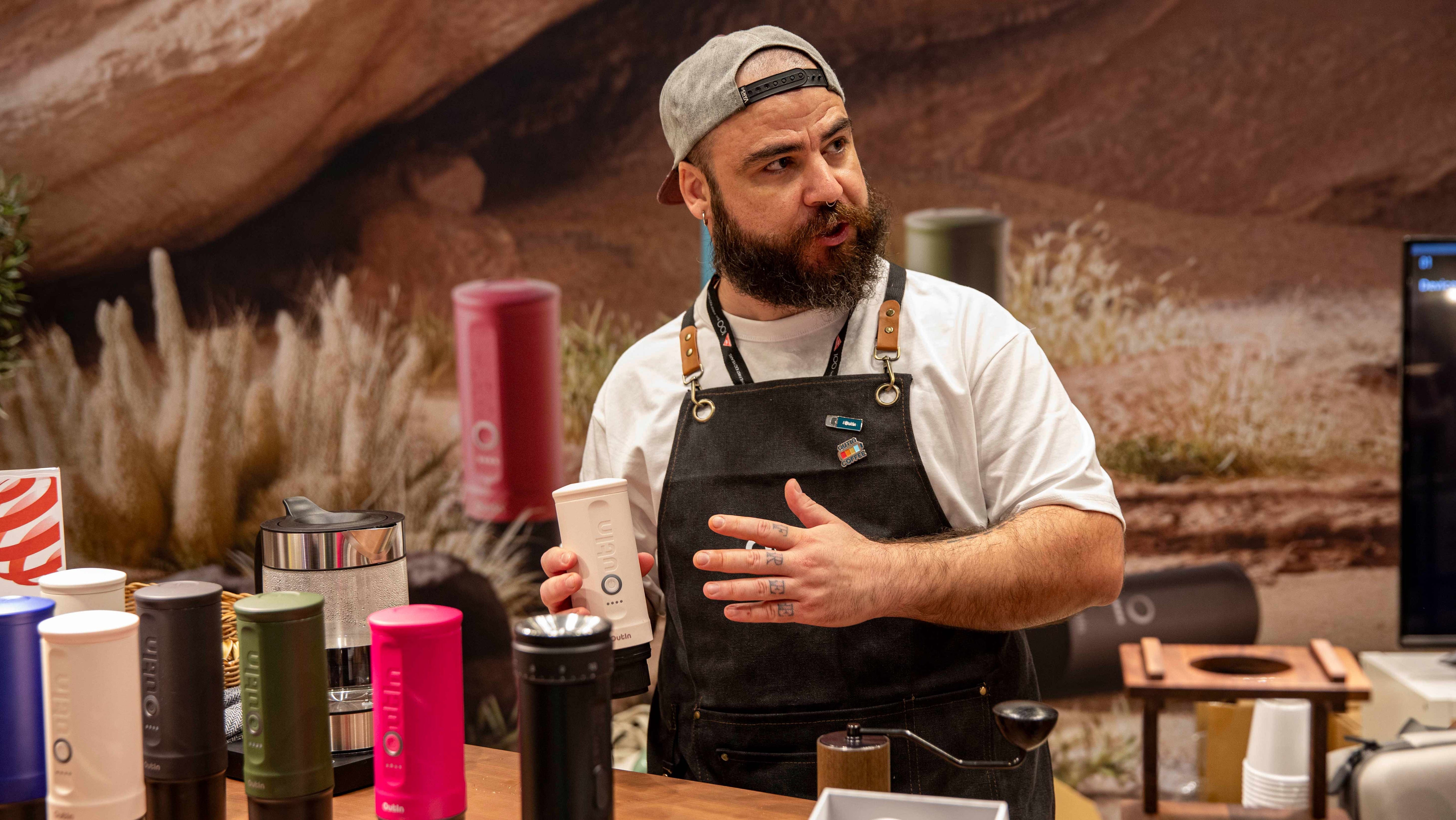 "Professional barista Matteo D'Ottavio demonstrates OutIn portable espresso machines while wearing a black apron, white t-shirt and backwards cap. Several colorful OutIn coffee devices are displayed on the wooden table in front of him.