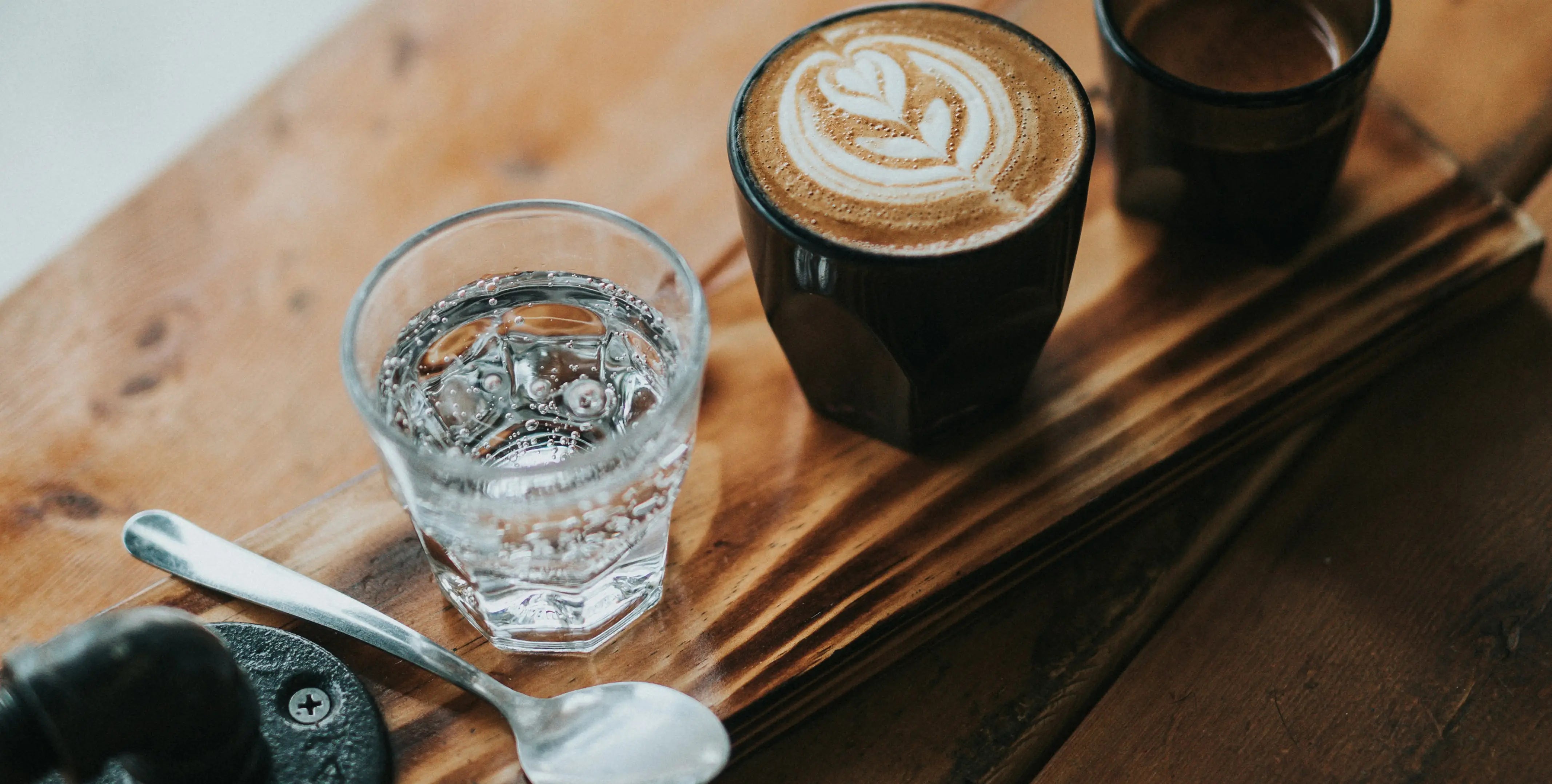 Latte Art mit Herz, dazu ein Glas Wasser und Löffel auf einem Holztablett