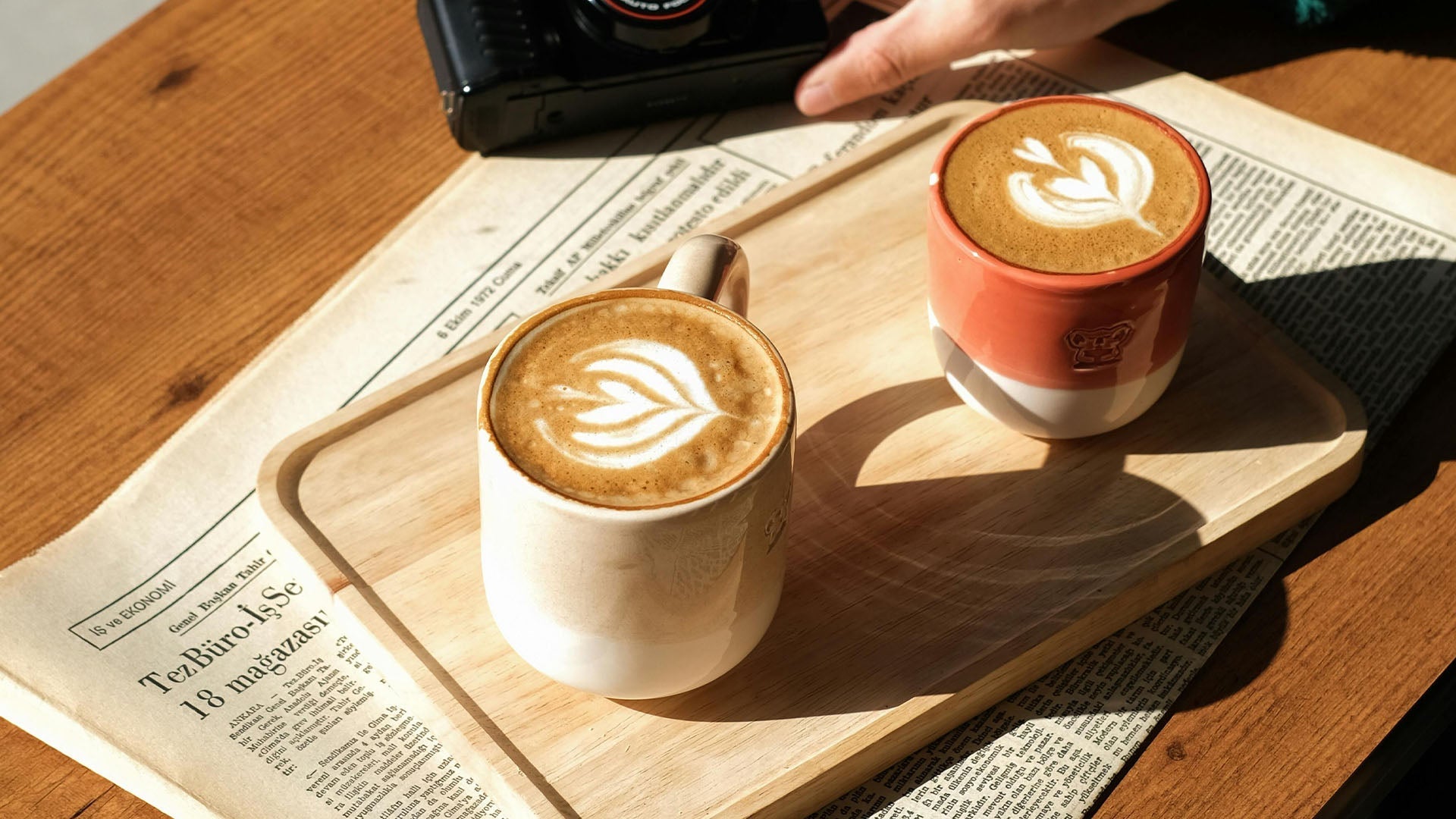 Zwei Kaffeetassen mit Latte-Art-Herzen auf Holzbrett im Sonnenlicht vor Zeitungshintergrund mit gemütlicher Café-Stimmung.