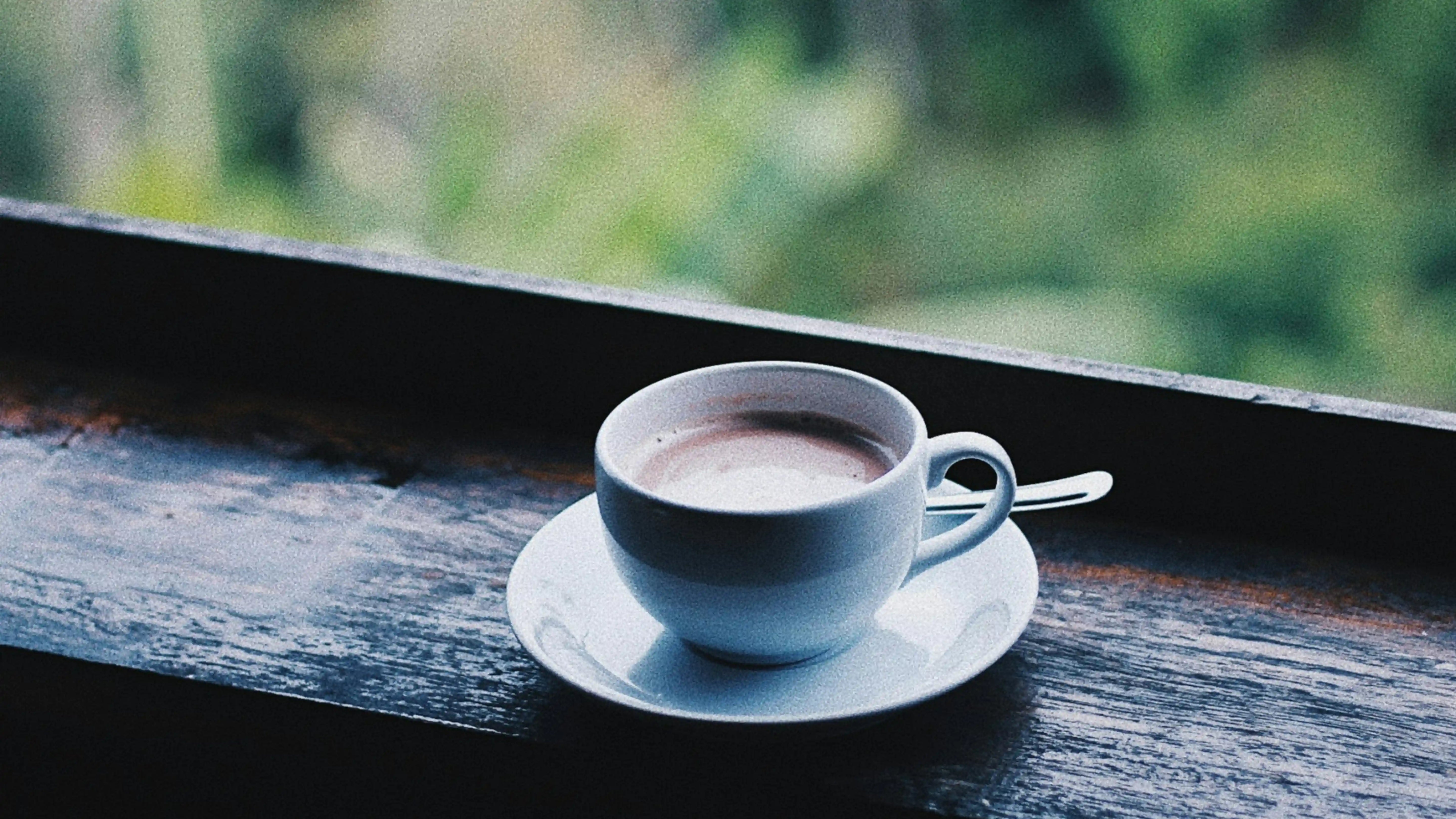 Cremiger Latte in weißer Keramiktasse mit Untertasse vor unscharfem, grünem Naturhintergrund – perfekte Morgenkaffee-Ästhetik