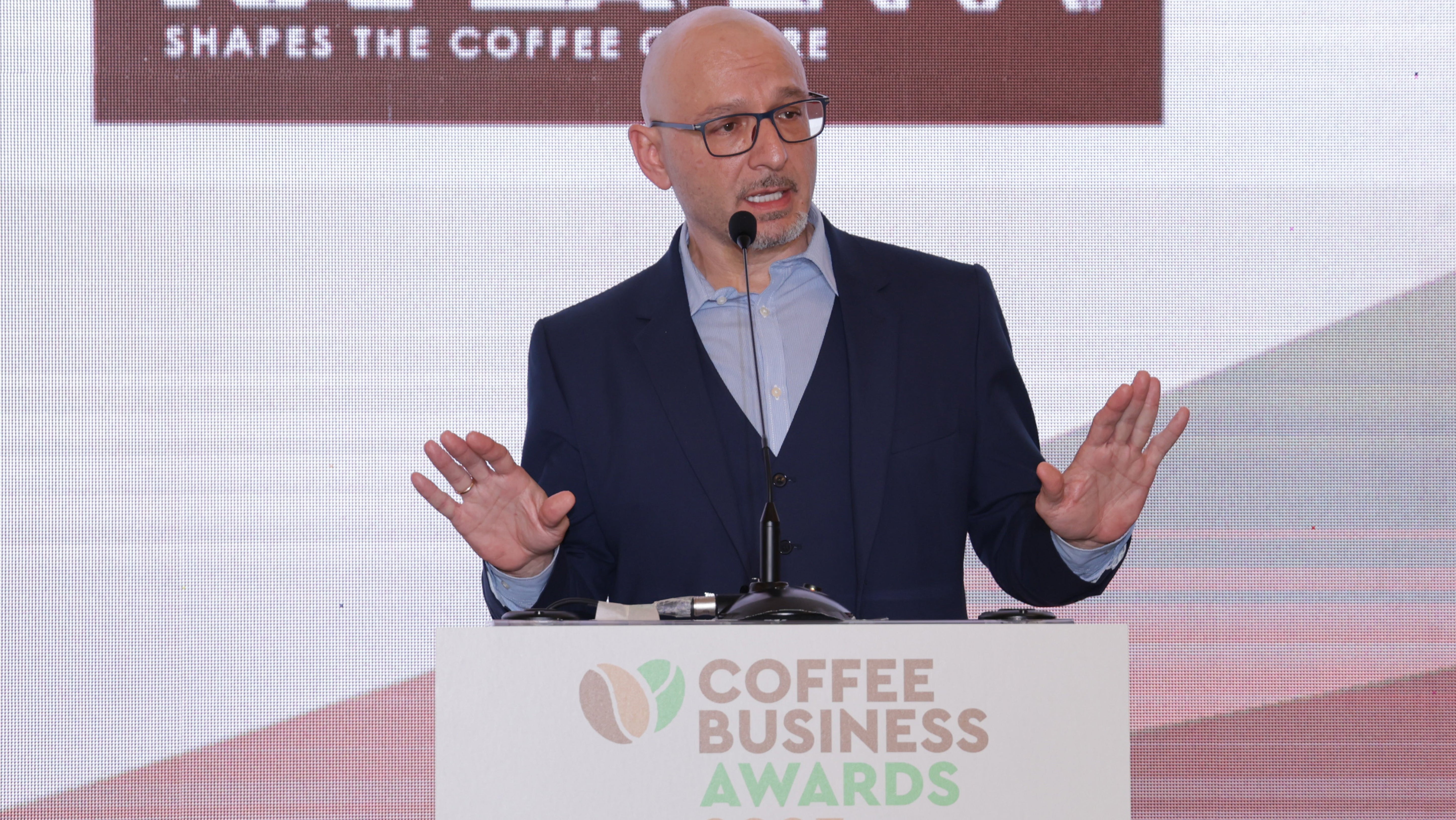 Panagiotis hält eine Rede bei den Coffee Business Awards. Der Mann im Anzug gestikuliert vor der Leinwand mit Kaffeemotto