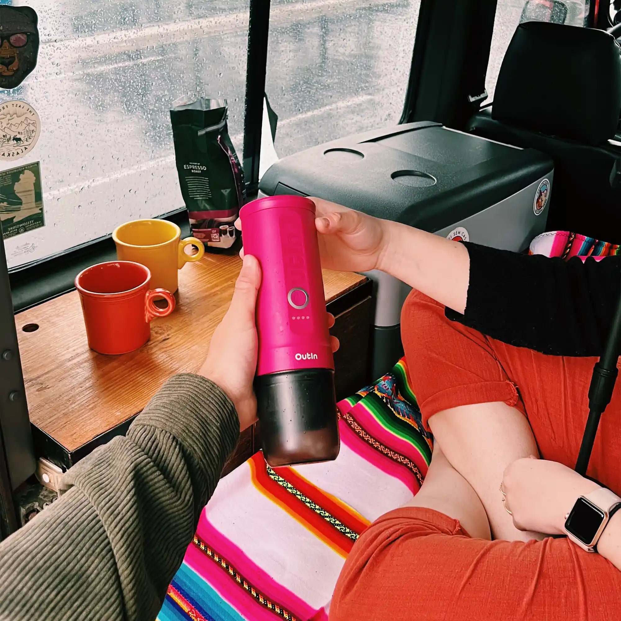 Pink OutIn Nano tragbare Espressomaschine in Händen, mit Tassen und Kaffeepulver auf Tisch im Auto, Regentropfen am Fenster.
