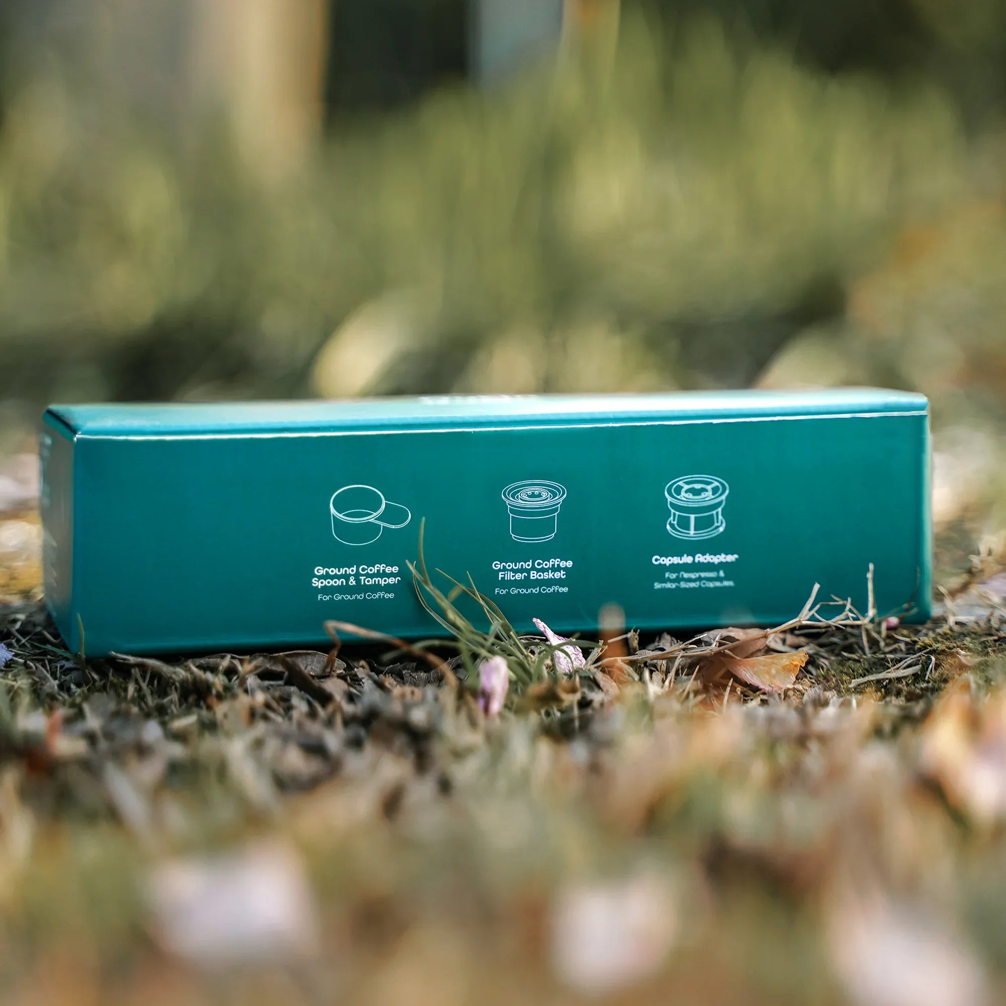 Outin teal box highlighting Ground Coffee Spoon, Filter Basket, and Capsule Adapter.