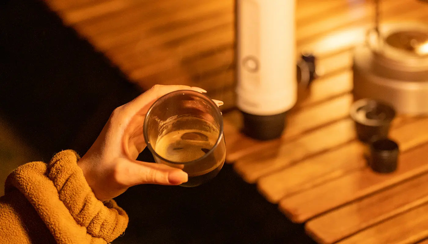 Person hält ein Glas Whisky in der Hand, im Hintergrund ein Tisch mit einer Flasche und weiteren Gläsern, warme Beleuchtung
