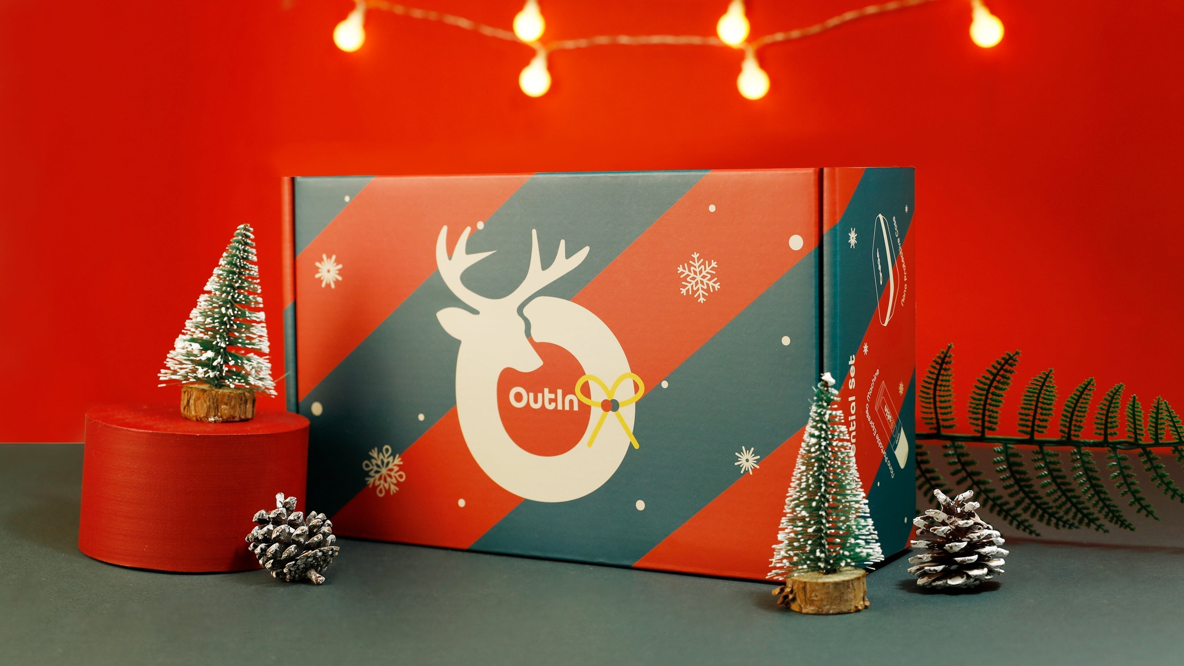 OutIn Nano schwarze tragbare Espressomaschine mit Weihnachtsgeschenkbox, Kaffeekapseln, Mini-Weihnachtsbaum und Lichterkette auf rotem Hintergrund