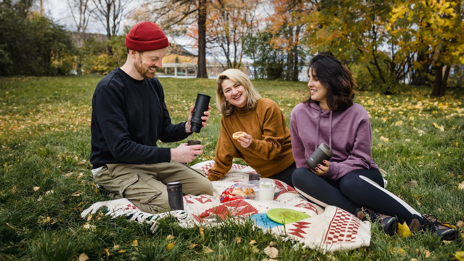 Eine Gruppe beim Picknick im Park, schwarze tragbare Kaffeemaschinen in der Hand, während sie den Herbst im Freien genießen