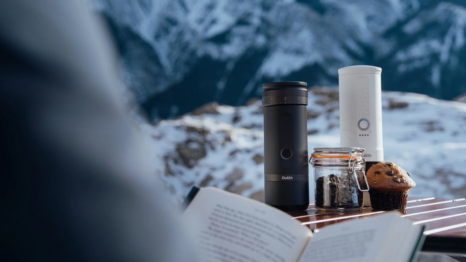 Auf einem Holztisch stehen zwei tragbare Espressomaschinen, im Vordergrund liegt ein offenes Buch und im Hintergrund sind schneebedeckte Berge zu sehen.