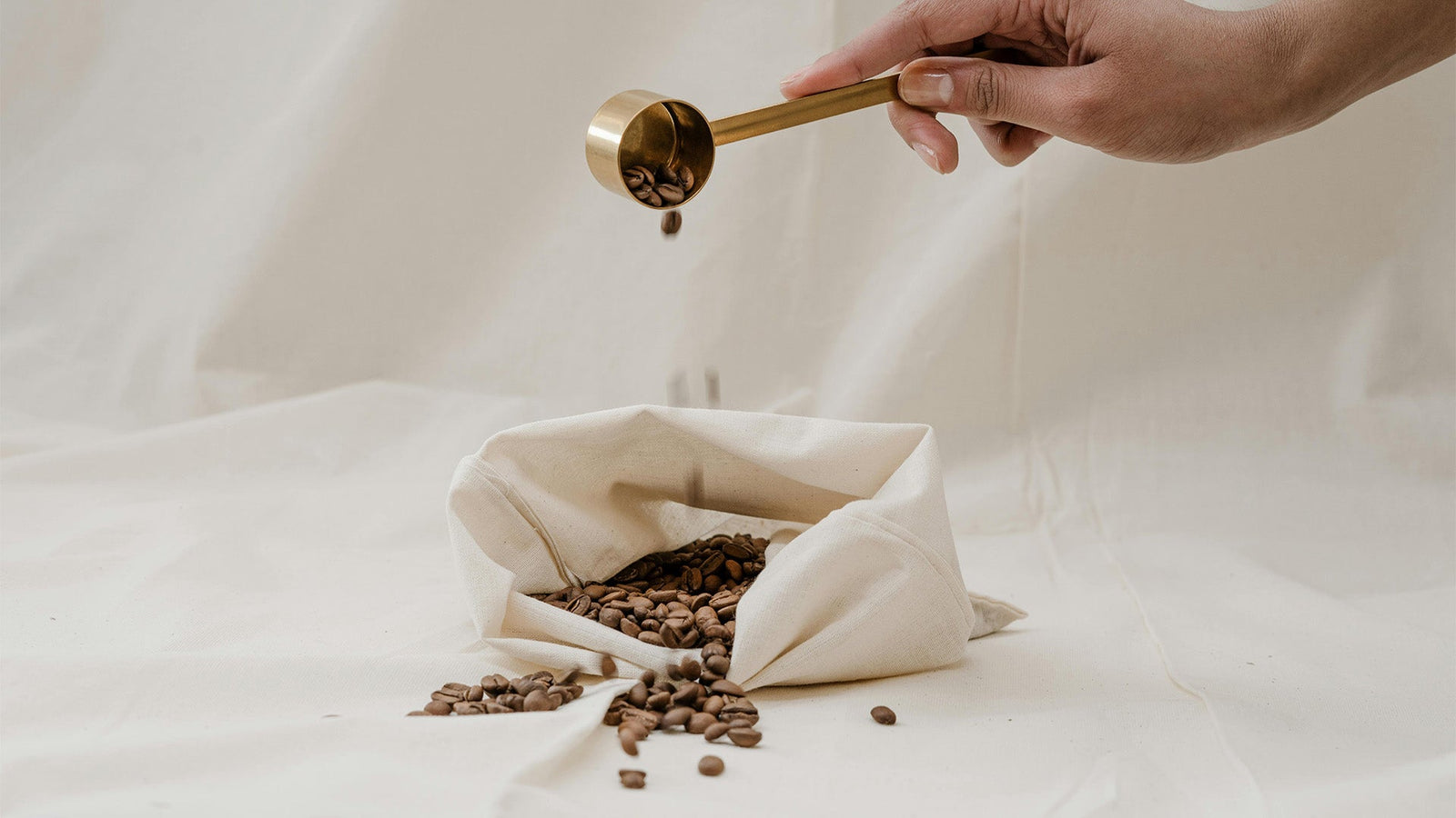 Hand mit Messlöffel aus Messing, die frische Kaffeebohnen aus einem weißen Stoffbeutel abmisst – minimalistisches Setup für die Kaffeezubereitung vor weißem Hintergrund.﻿