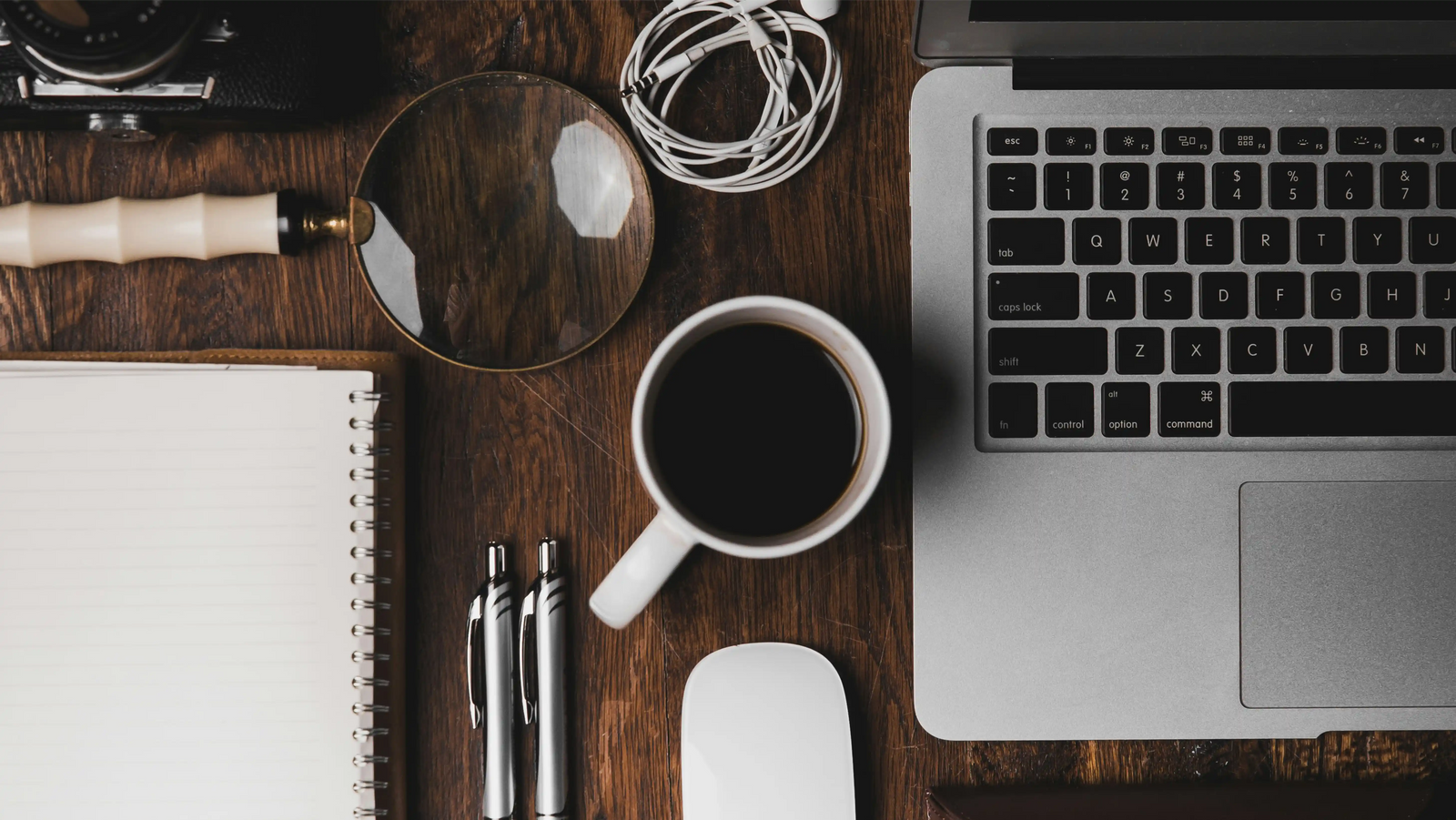 Minimalistisches Schreibtisch-Setup mit Laptop, Kaffeetasse, weißem Spiralblock, Lupe, weißen Kopfhörern und Stiften auf einer Holzoberfläche, aufgenommen aus der Vogelperspektive bei stimmungsvollem Licht