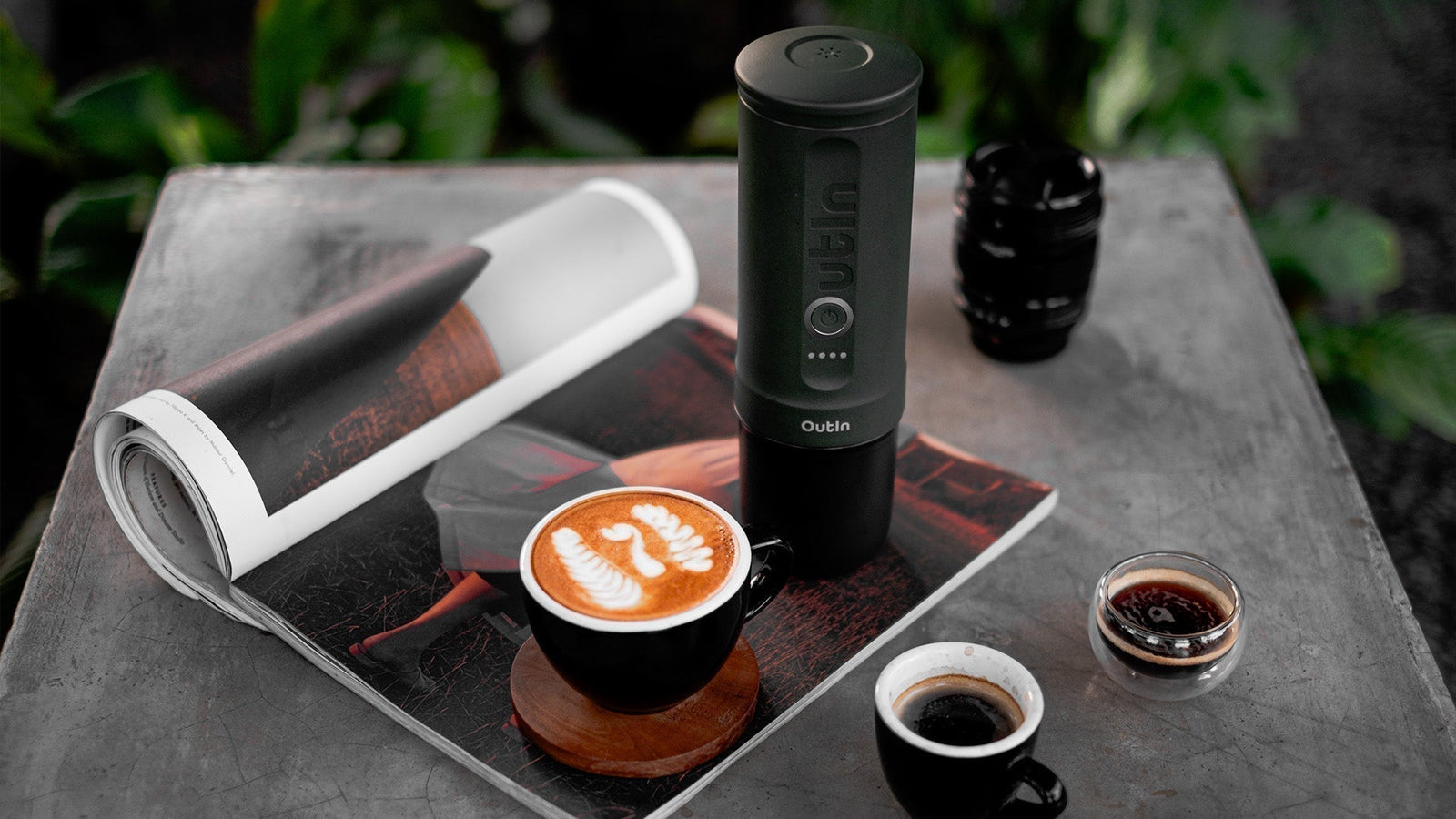Outin tragbare Espressomaschine mit Latte-Art-Kaffeetasse, Zeitschrift und Espresso-Shots auf Betontisch, die leistungsstarke Kaffeezubereitungsoptionen für Koffeinliebhaber präsentiert.