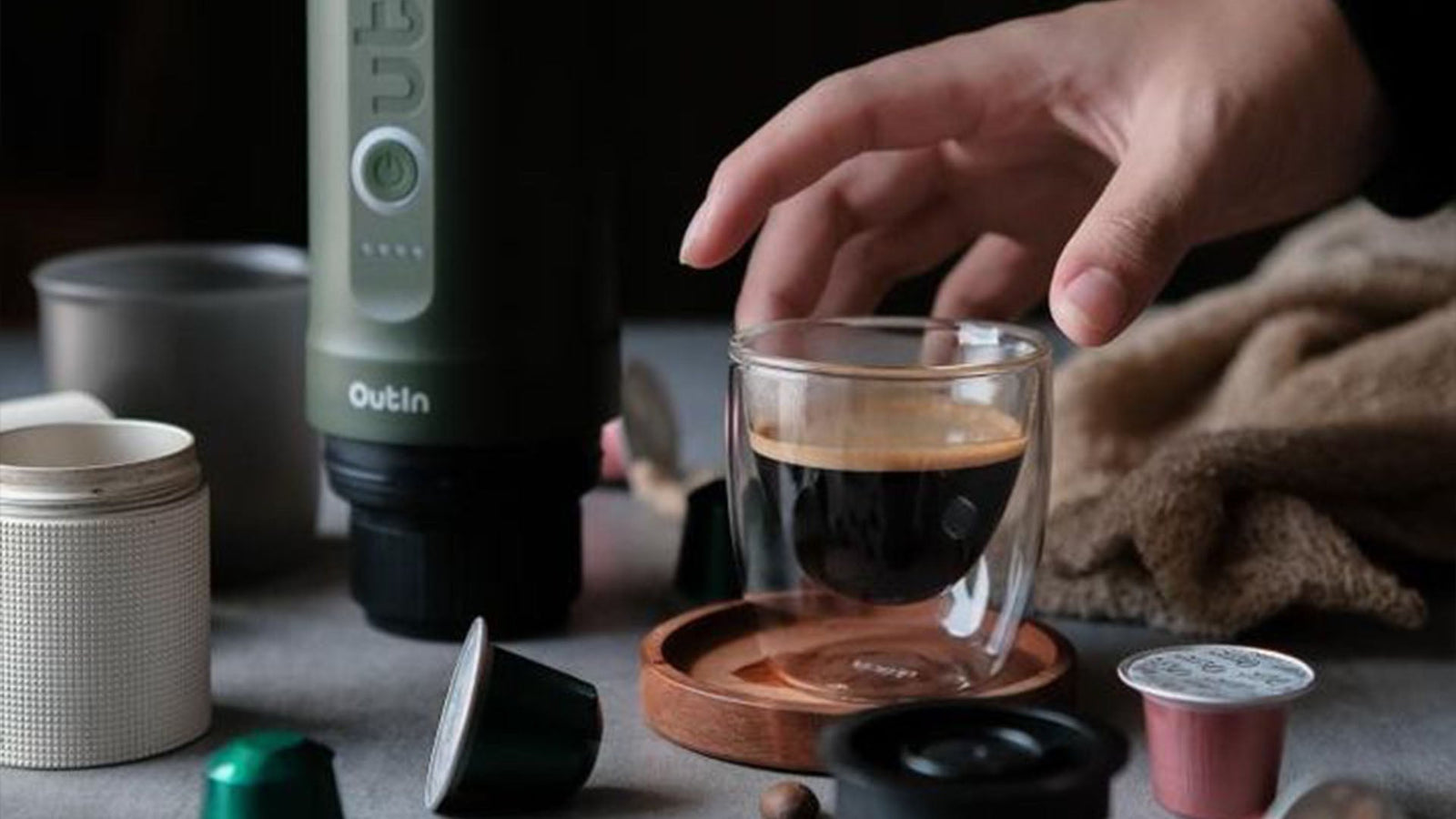 Eine Hand greift nach einem doppelwandigen Espresso-Glas neben einer tragbaren Outln-Kaffeemaschine und demonstriert praktische Kaffeealternativen für zuhause im Vergleich zu Instantkaffee – ideal für die Zubereitung unterwegs