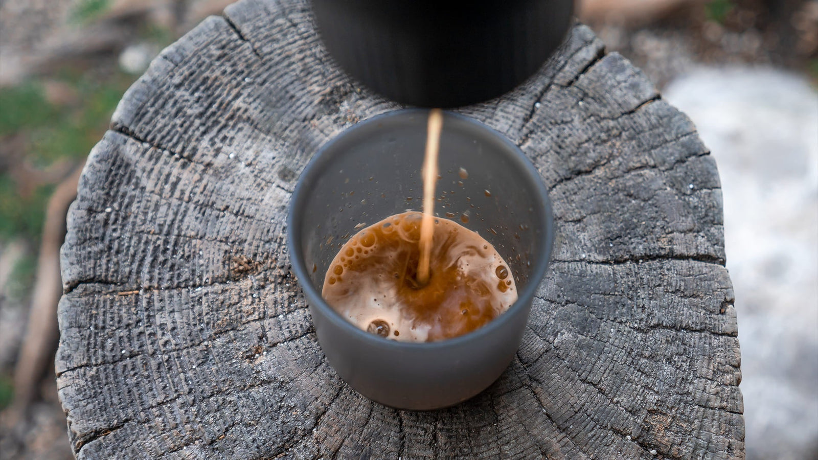 Frisch gebrühter Kaffee wird in eine graue Tasse auf einem Baumstumpf gegossen, wobei die satte Bernsteinfarbe und die Crema während des Brühvorgangs im Freien sichtbar werden